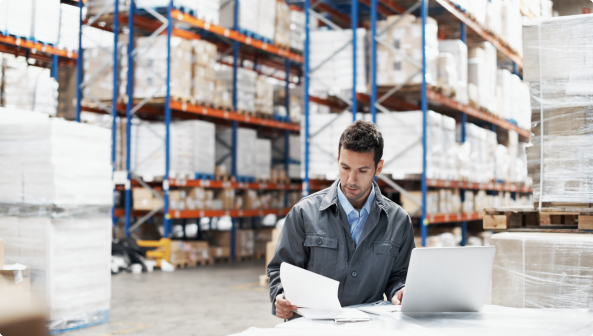 Male looking at a paper and working on a laptop in a warehouse